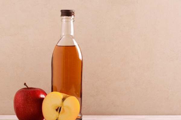 A bottle of apple cider vinegar is placed next to fresh apples on a neutral background.