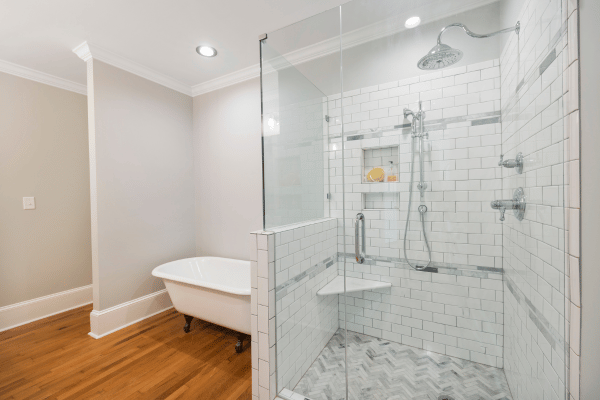 A modern bathroom featuring a glass-enclosed shower with white subway tiles, a rainfall showerhead, and a freestanding clawfoot bathtub on warm wooden flooring.