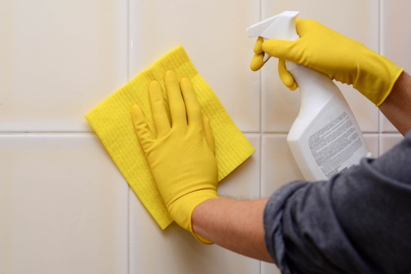 Person wearing yellow gloves scrubbing shower tiles with a yellow cloth and spray bottle.